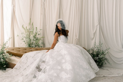 Woman in a white wedding dress sitting on a couch with floral decorations and white curtains in the background.