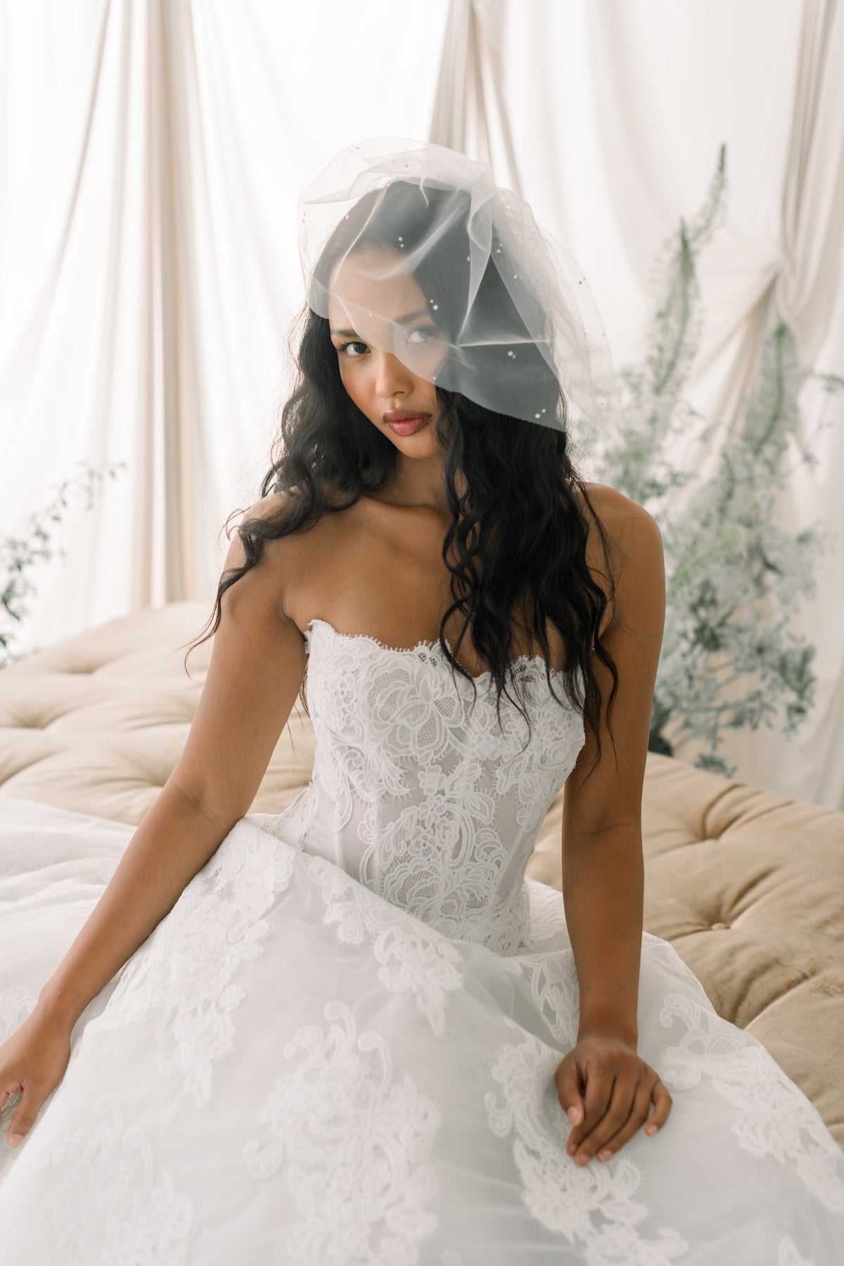 Woman in a white lace wedding dress with a veil sitting on a couch.