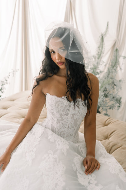 Woman in a white lace wedding dress with a veil sitting on a couch.