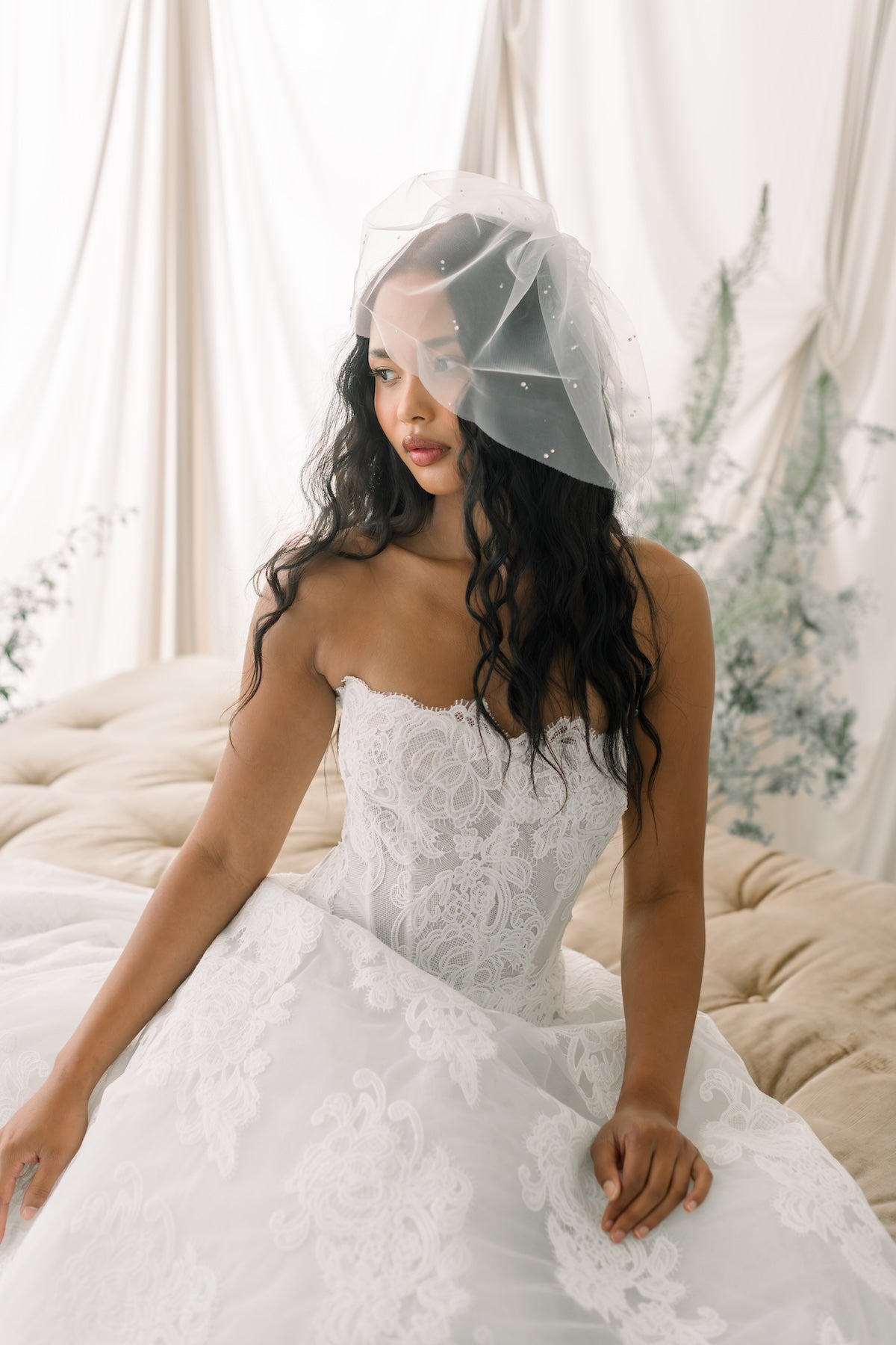 Woman in a white lace wedding dress with a veil sitting on a couch.