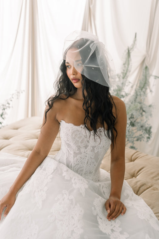 Woman in a white lace wedding dress with a veil sitting on a couch.
