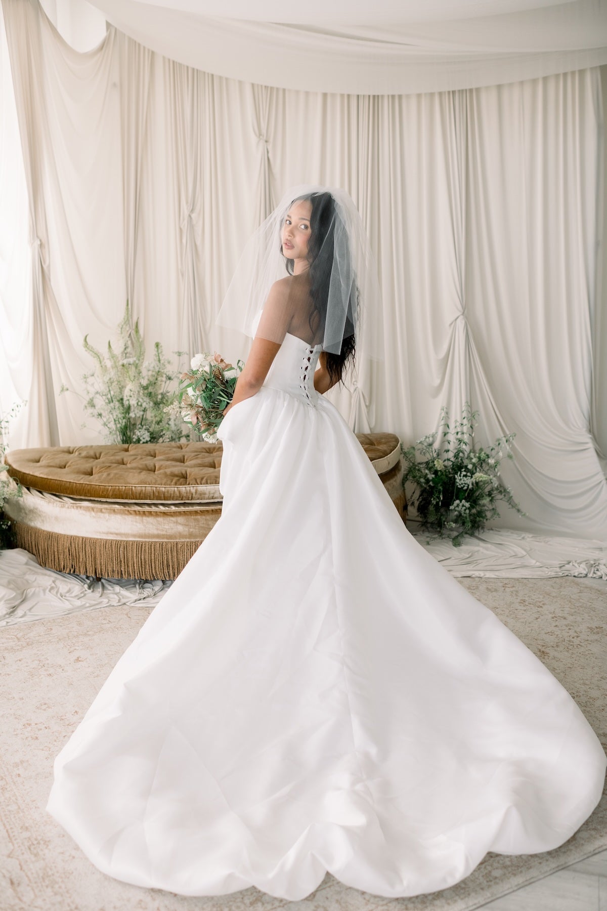 Woman in a white wedding dress standing in a decorated room with floral arrangements and draped curtains.