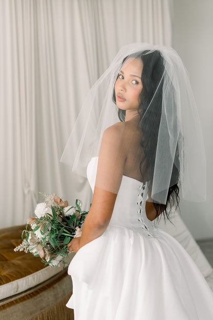 Woman in a white wedding dress with a veil, holding a bouquet, in a softly lit room.
