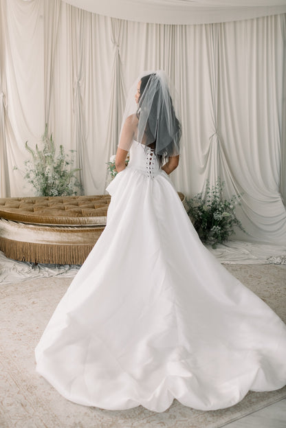 Bride in a white wedding dress with a long train standing in front of a draped white curtain.