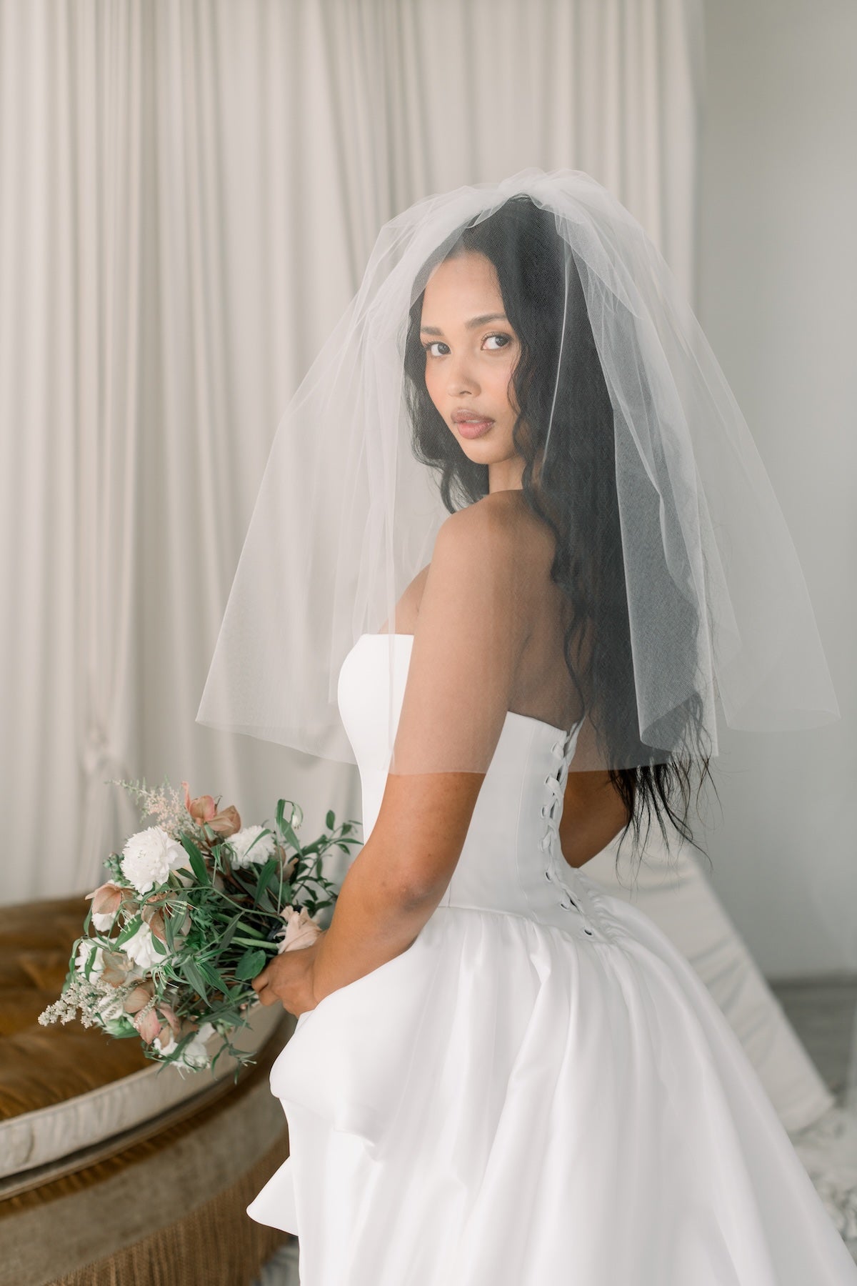 Bride in a white dress with a veil holding a bouquet indoors.