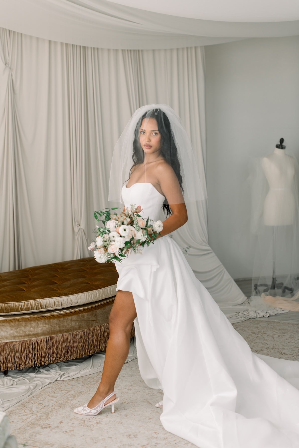 Bride in a white wedding dress holding a bouquet in a room with curtains and a mannequin.