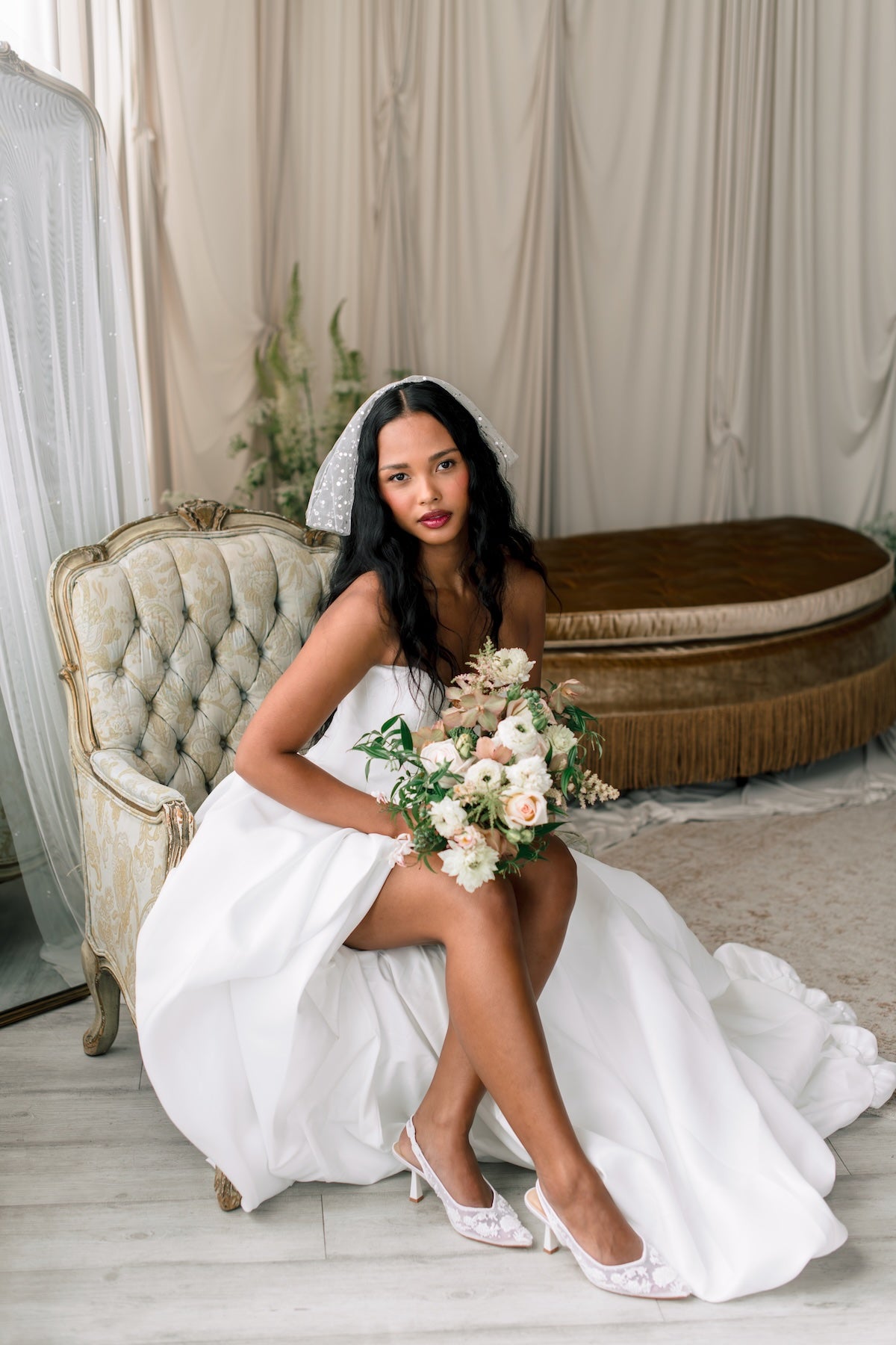 Bride in a white dress sitting on a vintage chair holding a bouquet of flowers.