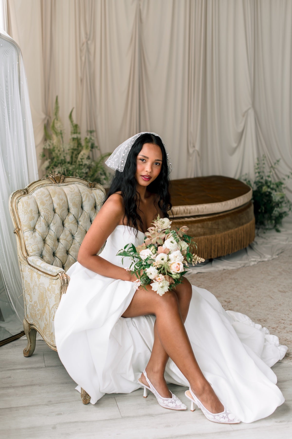 Bride in a white gown holding a bouquet in a decorated room with curtains and furniture.