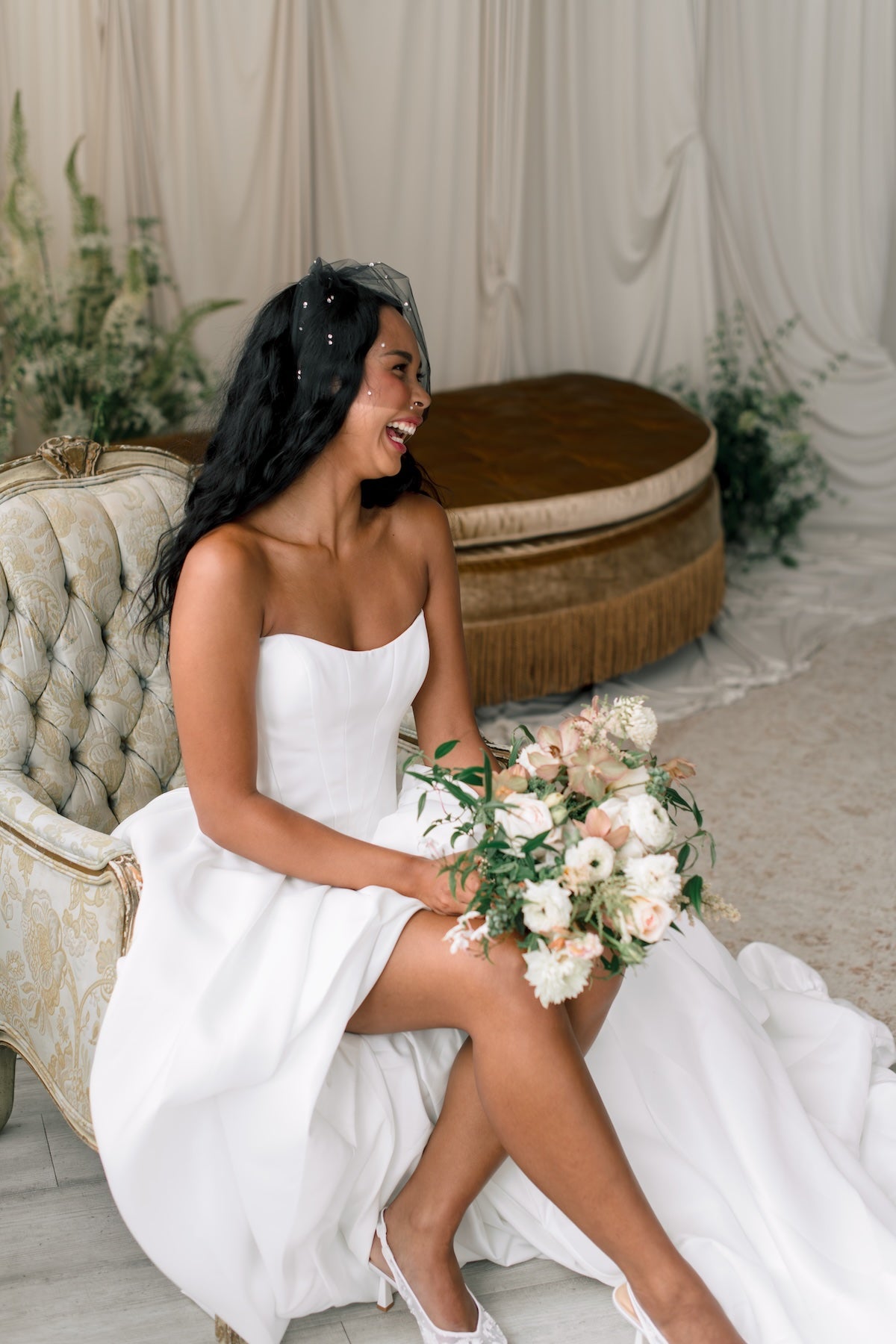 Bride in a white dress holding a bouquet, sitting on a decorative chair with a neutral background.