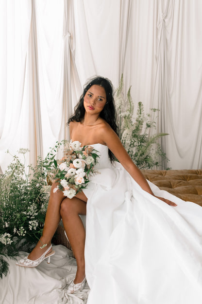 Woman in a white wedding dress holding a bouquet of flowers, sitting on a couch with a floral backdrop.