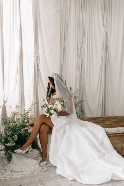 Bride in a white gown with a long veil sitting against a draped curtain backdrop.