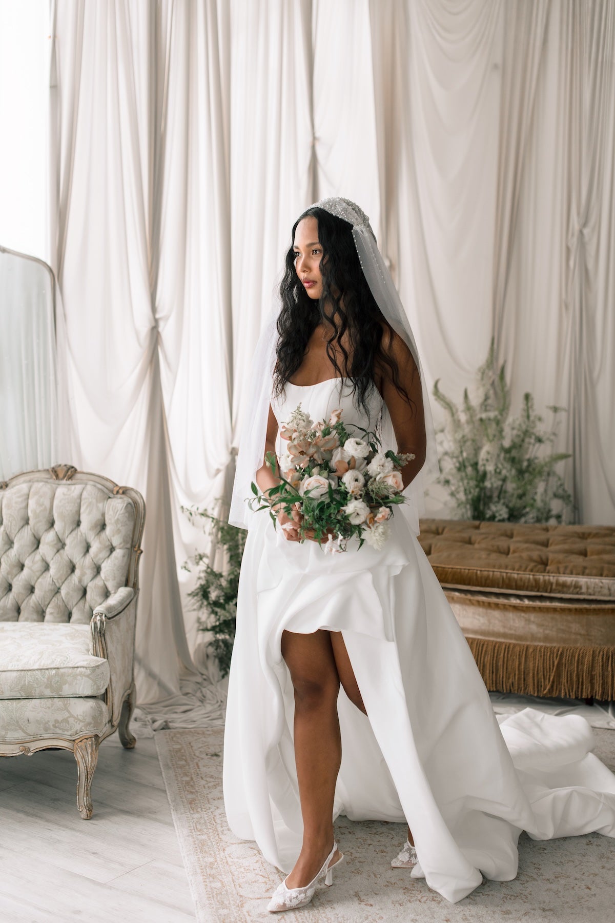 Bride in a white wedding dress holding a bouquet in a decorated room with curtains and furniture.