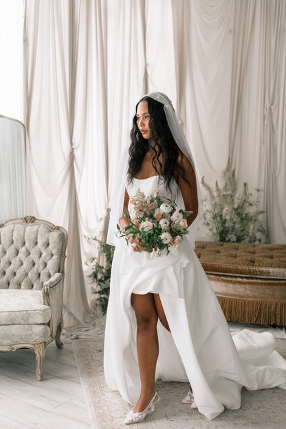 Bride in a white wedding dress holding a bouquet in a decorated room with curtains and furniture.