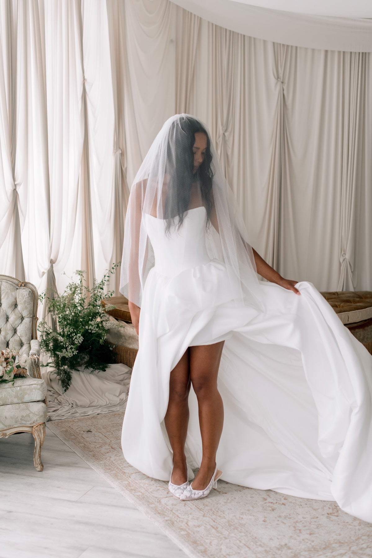 Woman in a white wedding dress with a veil standing in a room with curtains and a chair.