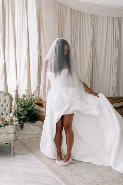 Woman in a white wedding dress with a veil standing in a room with curtains and a chair.