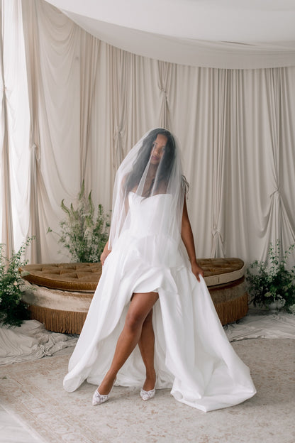 Bride in a white wedding dress with a long veil standing in a softly lit room with draped curtains and plants.