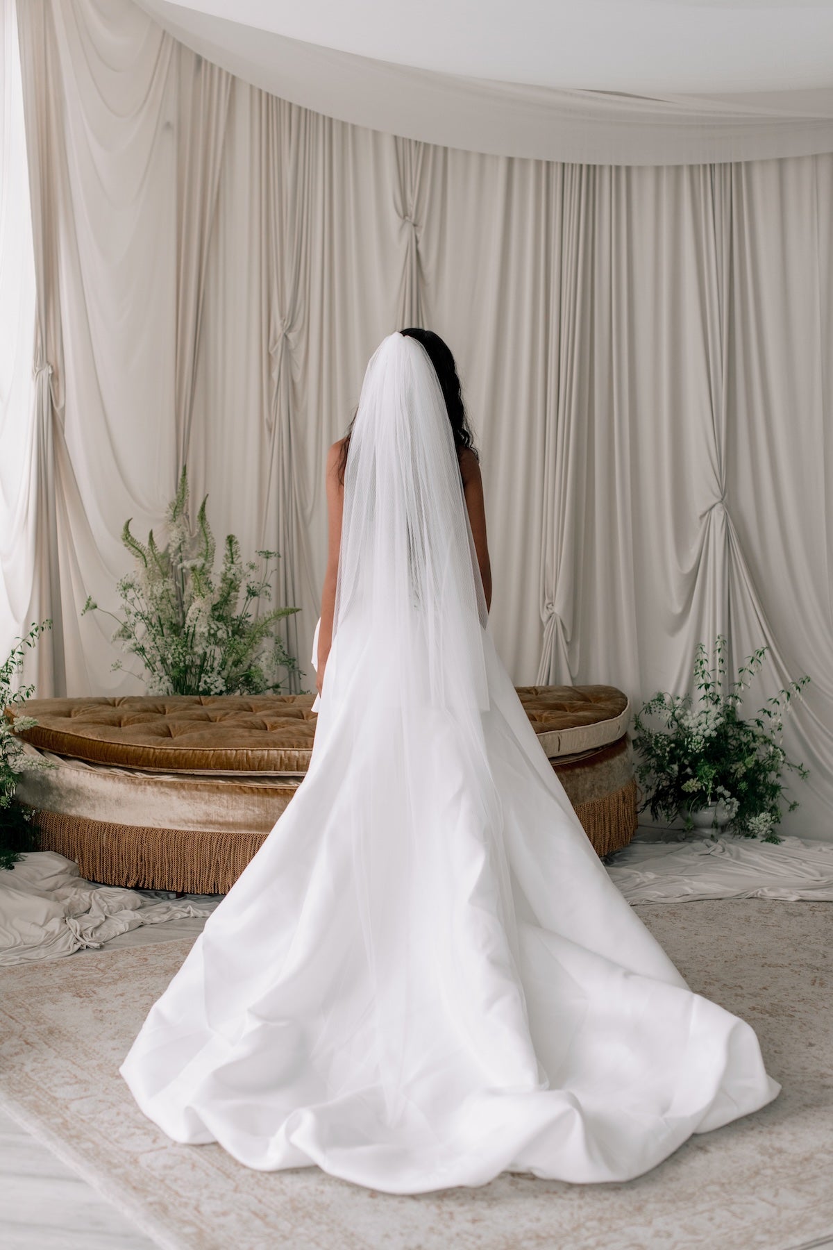 Person wearing a long white wedding veil in a decorated indoor setting with curtains and plants.