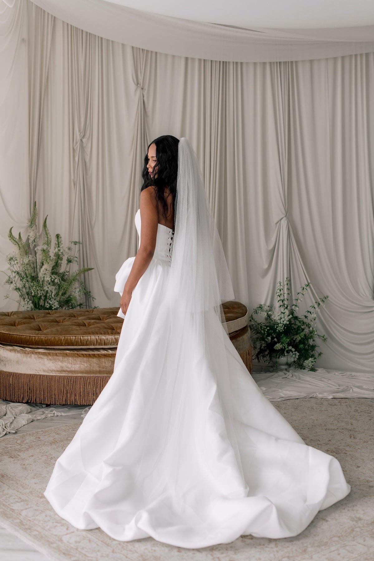 Woman in a white wedding dress with a long veil standing in front of draped curtains.