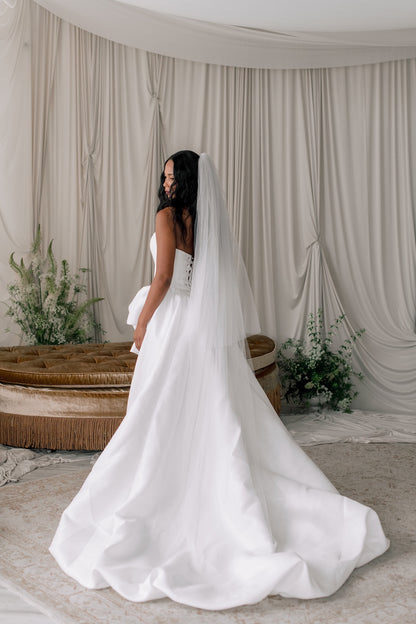 Woman in a white wedding dress with a long veil standing in front of draped curtains.