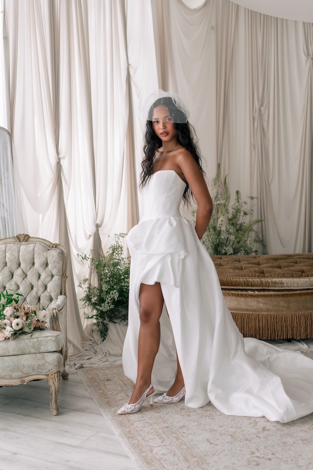 Woman in a white wedding dress standing in a decorated room with curtains and floral arrangements.
