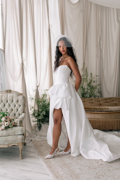 Woman in a white wedding dress standing in a decorated room with curtains and floral arrangements.