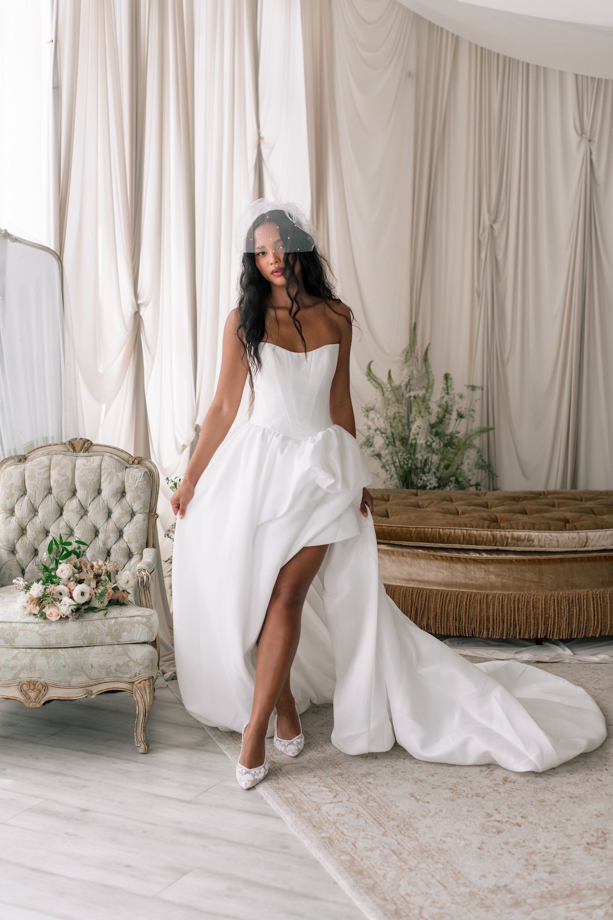 Woman in a white wedding dress standing in a decorated room with curtains and floral arrangements.
