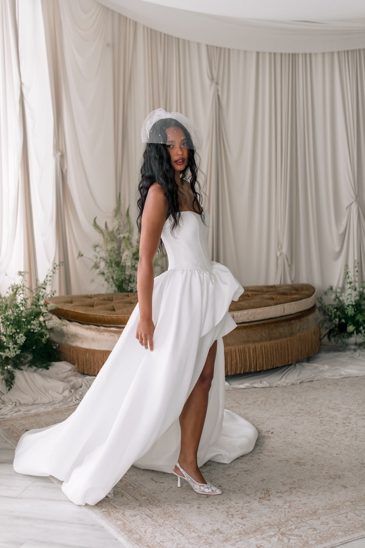 Woman in a white wedding dress standing in a decorated room with curtains and plants.