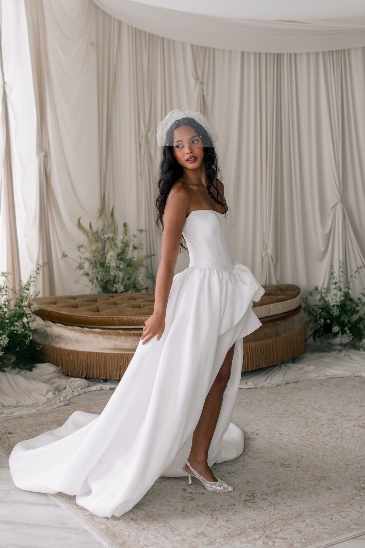 Woman in a white wedding dress standing in a decorated indoor setting with draped curtains and plants.