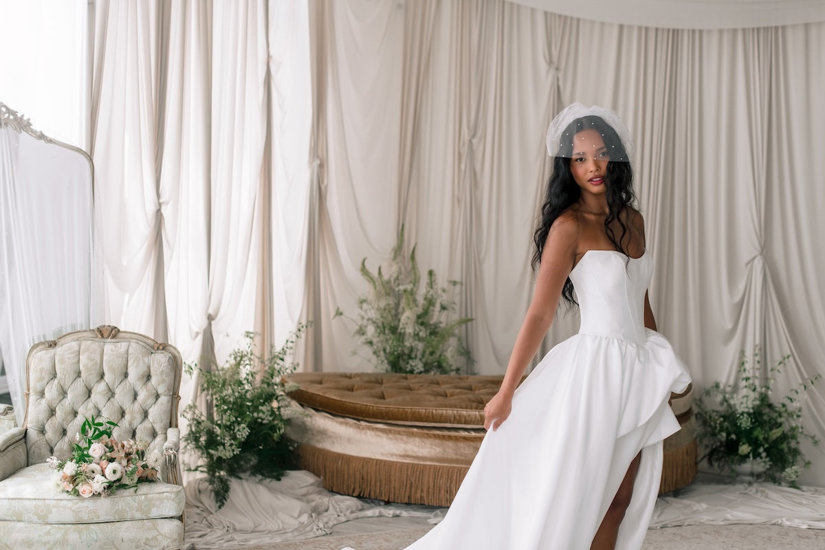Woman in a white wedding dress standing in a decorated room with floral arrangements and draped curtains.