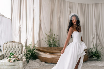 Woman in a white wedding dress standing in a decorated room with floral arrangements and draped curtains.