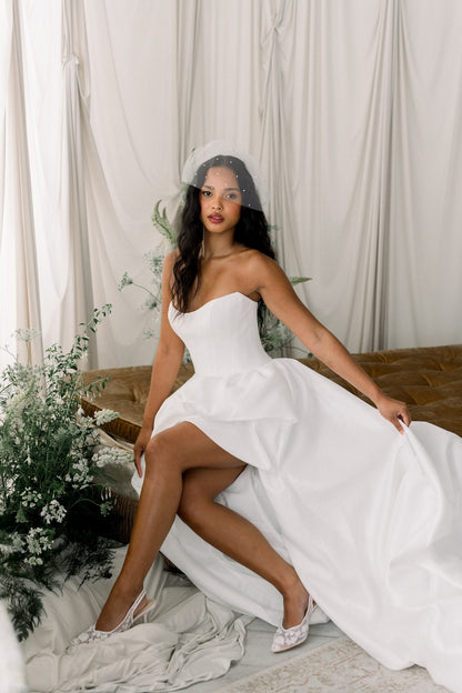 Woman in a white strapless dress with a high slit, sitting on a wooden floor with white curtains and plants in the background.