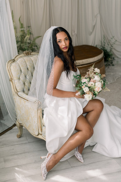 Bride in a white dress with a veil holding a bouquet, sitting on a chair in a decorated room.
