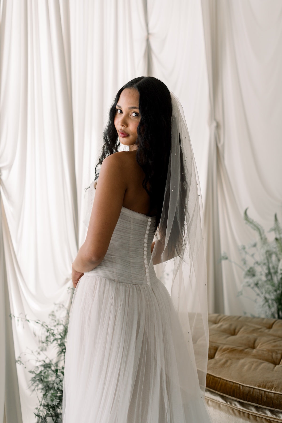 Woman in a white wedding dress with a veil standing in front of white curtains.