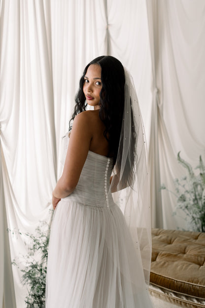 Woman in a white wedding dress with a veil standing in front of white curtains.
